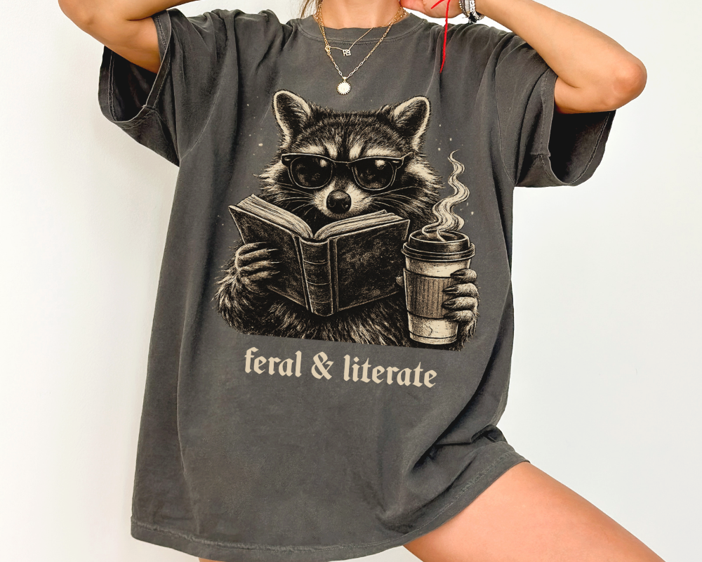 Vintage raccoon reading a book while holding a coffee cup with the phrase Feral & Literate printed on a Comfort Colors pepper t-shirt.