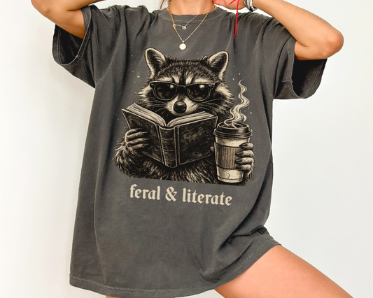 Vintage raccoon reading a book while holding a coffee cup with the phrase Feral & Literate printed on a Comfort Colors pepper t-shirt.