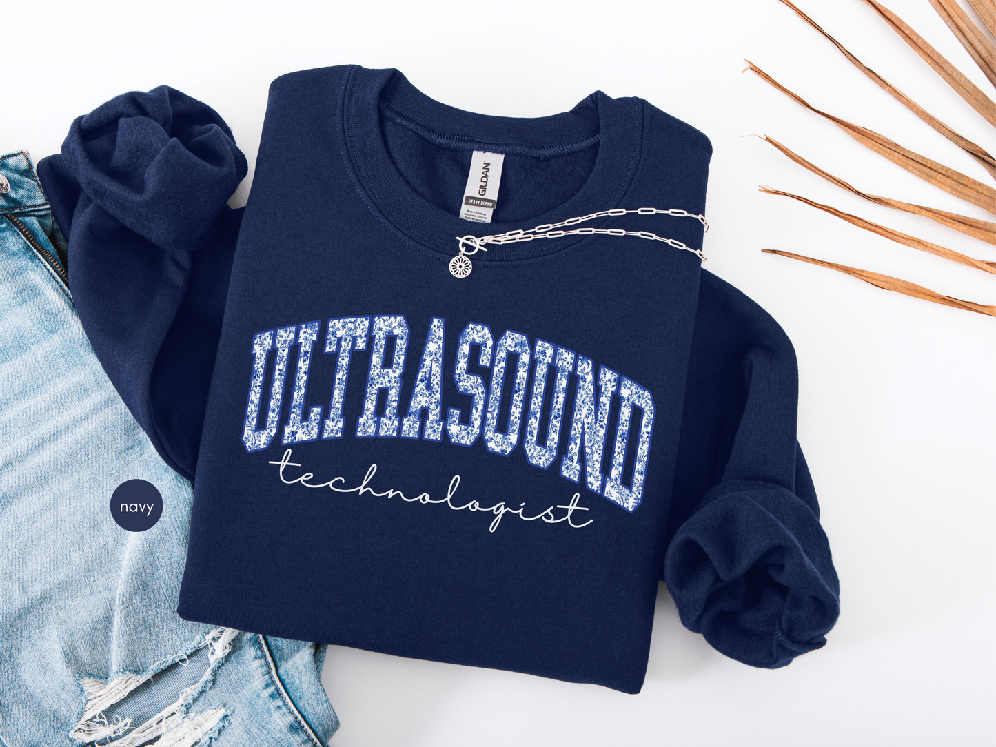 Navy blue Ultrasound Technologist sweatshirt with collegiate-style lettering, folded flat with denim jeans and necklace for medical professional gift