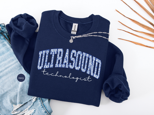 Navy blue Ultrasound Technologist sweatshirt with collegiate-style lettering, folded flat with denim jeans and necklace for medical professional gift