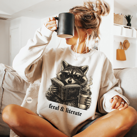 Woman wearing a sand Feral and Literate sweatshirt featuring a vintage raccoon in sunglasses reading a book and holding a cup of coffee.