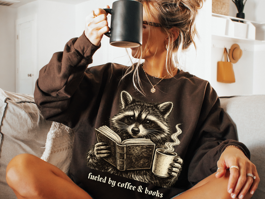 Woman wearing a chocolate Fueled by Coffee and Books sweatshirt featuring a vintage raccoon reading a book and holding a steaming cup of coffee.