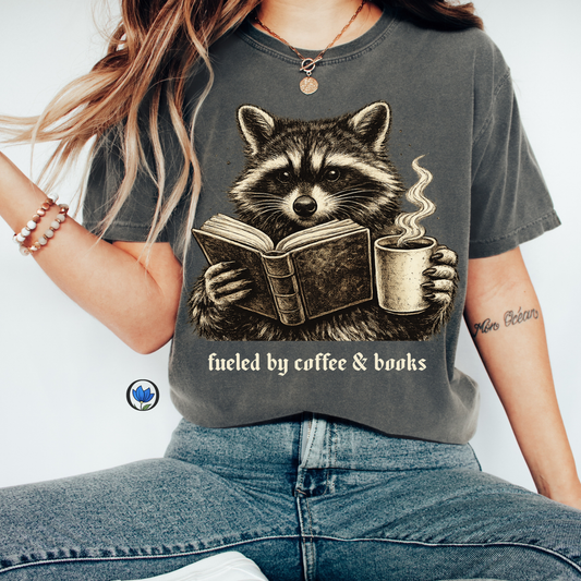 Vintage raccoon reading a book while holding a hot cup of coffee with the phrase "Fueled by Coffee & Books" on a Comfort Colors graphic t-shirt.